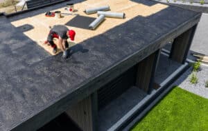 A construction worker is installing EPDM roofing material on a contemporary home under clear skies, surrounded by tools and prepared materials for the roofing project.